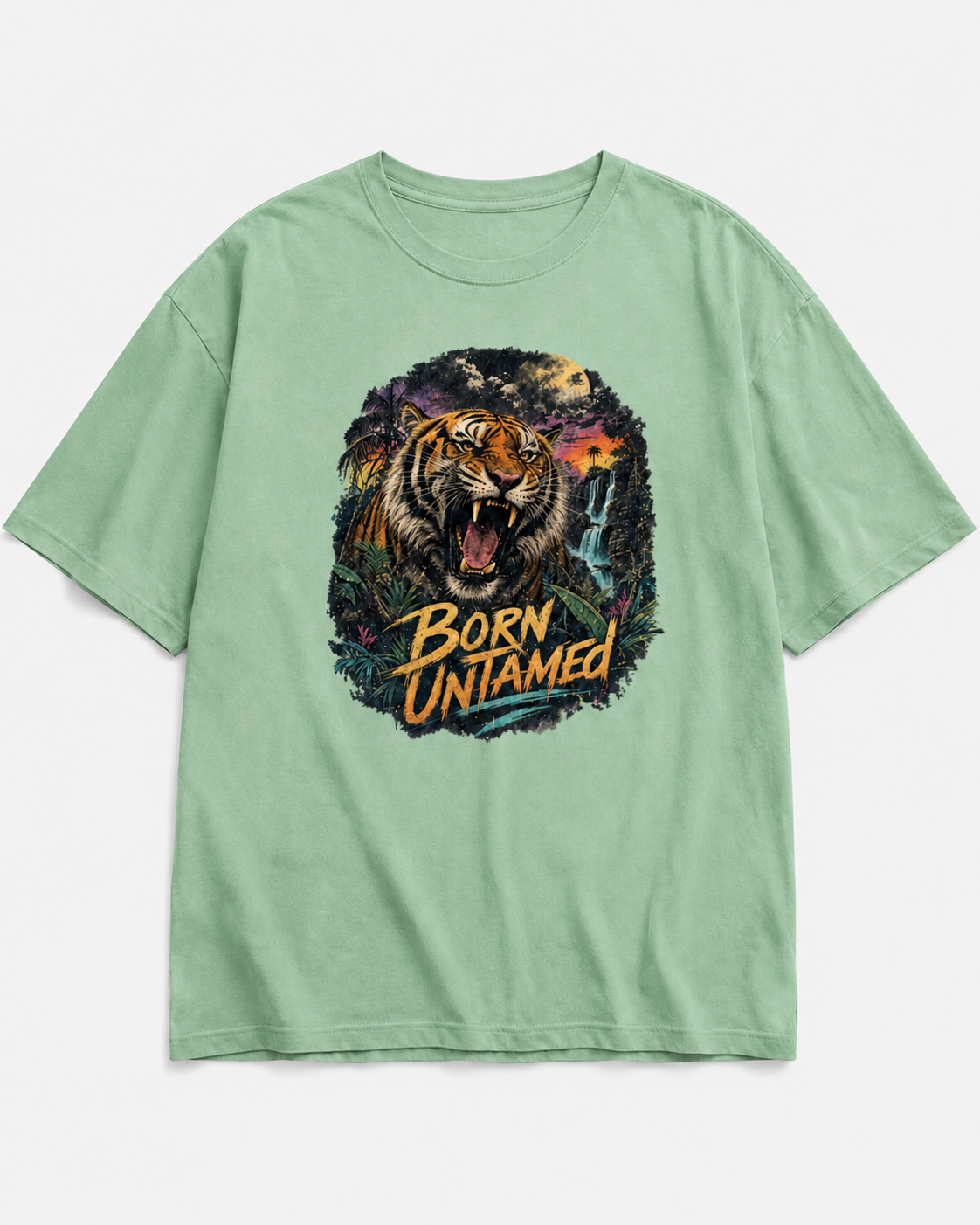 A light sage green short-sleeve t-shirt with a large graphic print of a roaring tiger and the text 'BORN UNTAMED' on the front, displayed flat against a light gray background.