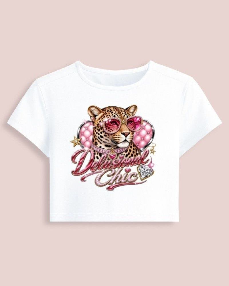 A white short-sleeve crop top featuring a graphic of a leopard wearing pink heart-shaped sunglasses with the text 'Delicious Chic' in pink script, displayed on a solid pale pink background.
