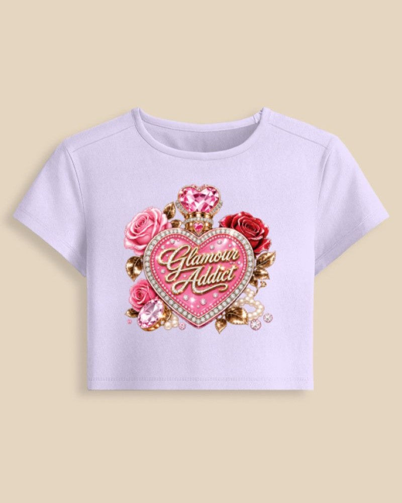 A light lavender short-sleeve crop top featuring a large graphic on the front with pink and red roses, a heart-shaped gem, and the words 'Glamour Addict' in gold script, displayed against a solid beige background.