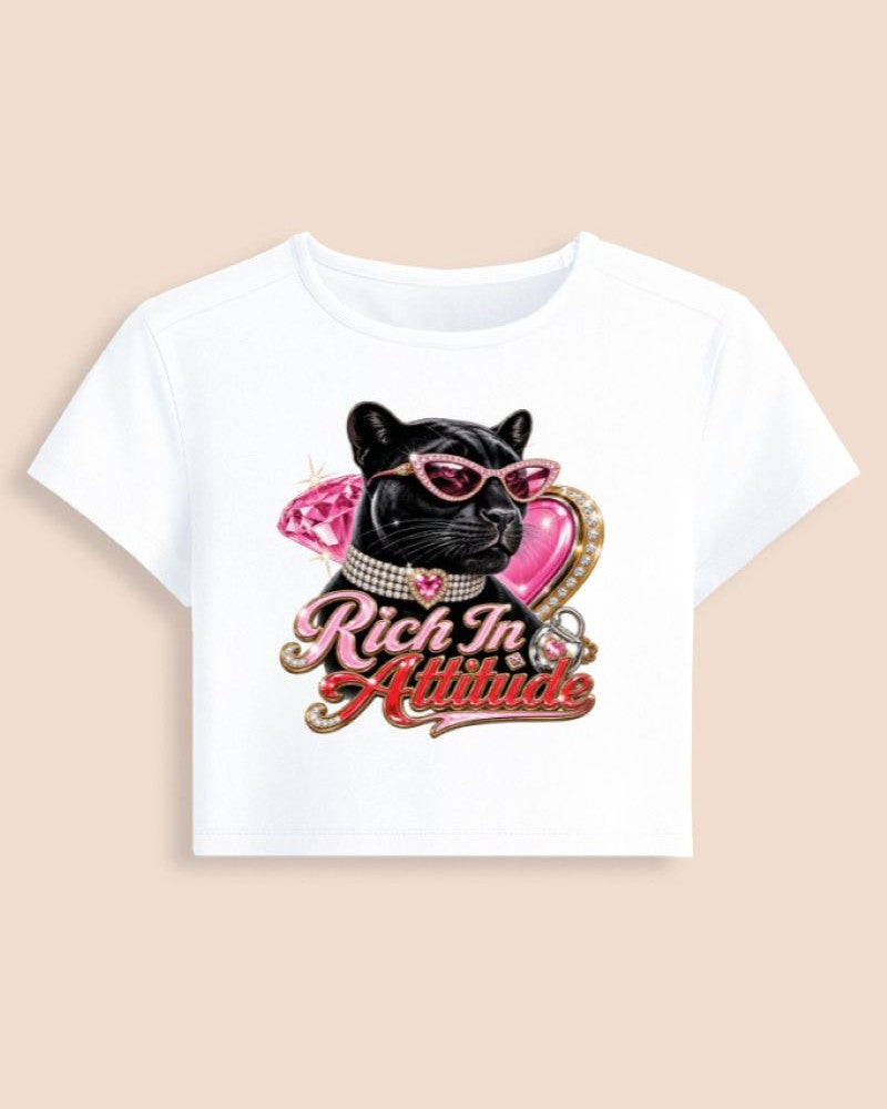 A white short-sleeve crop top featuring a graphic of a black cat wearing pink sunglasses and a pink bow, with the text 'Rich In Attitude' in pink and gold script, displayed on a solid beige background.