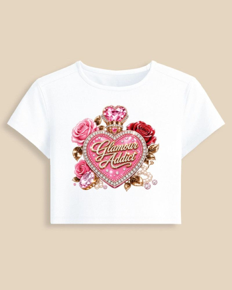 A white short-sleeve crop top t-shirt featuring a large graphic print of pink and red roses, a pink heart-shaped gem, and a central glittering pink heart with the words 'Glamour Addict' in gold script, displayed against a solid beige background.