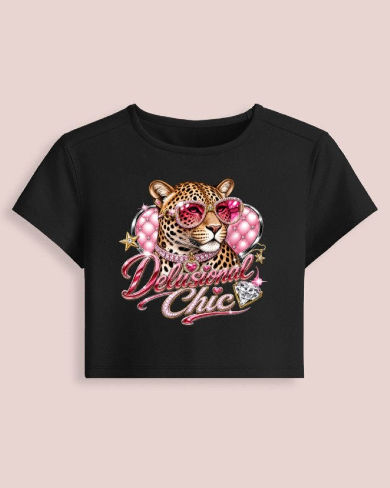 A black short-sleeve crop top t-shirt featuring a graphic of a leopard wearing pink heart-shaped sunglasses, surrounded by pink polka-dot hearts and the text 'Delicious Chic' in pink script, displayed against a solid pale pink background.