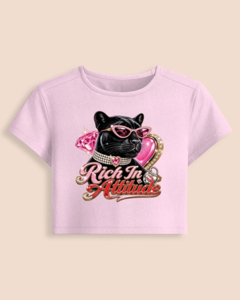 A light pink short-sleeve crop top featuring a graphic of a black cat wearing pink sunglasses and a pink heart, with the text 'Rich In Attitude' in a gold and pink script, displayed on a solid beige background.