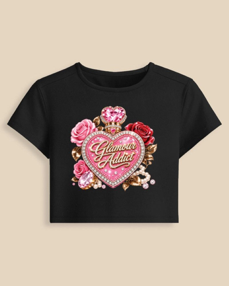 A black short-sleeve crop top t-shirt featuring a large graphic print on the front. The graphic displays a central pink heart with the words 'Glamour Addict' in gold script, surrounded by pink and red roses, gold leaves, and sparkling gem details, set against a solid beige background.