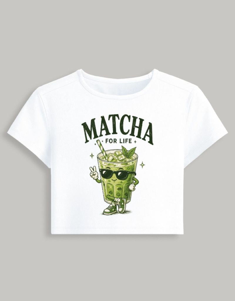 A white short-sleeve crop top t-shirt featuring a green cartoon matcha drink character wearing sunglasses and making a peace sign, with the text 'MATCHA FOR LIFE' printed above it, shown against a solid gray background.