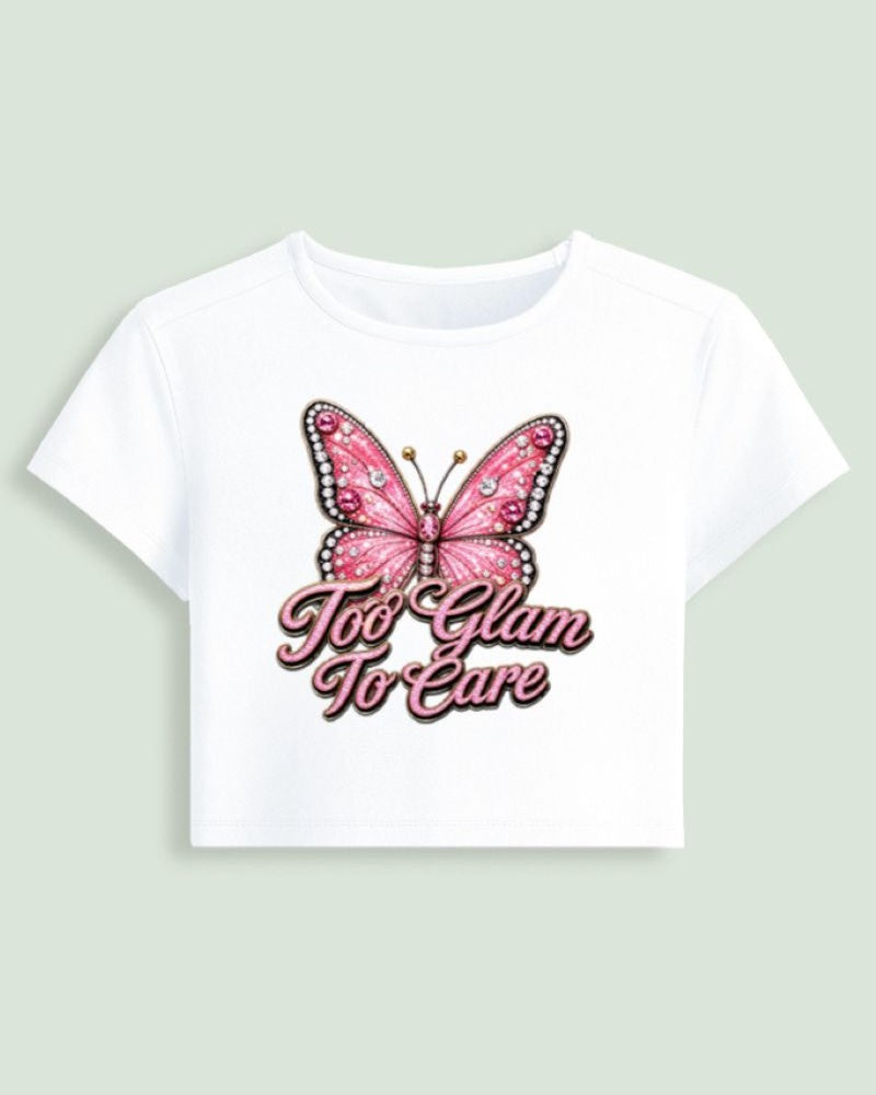 A white short-sleeve crop top featuring a graphic of a pink butterfly with rhinestone details and the text 'Too Glam To Care' in pink script, displayed against a light sage green background.