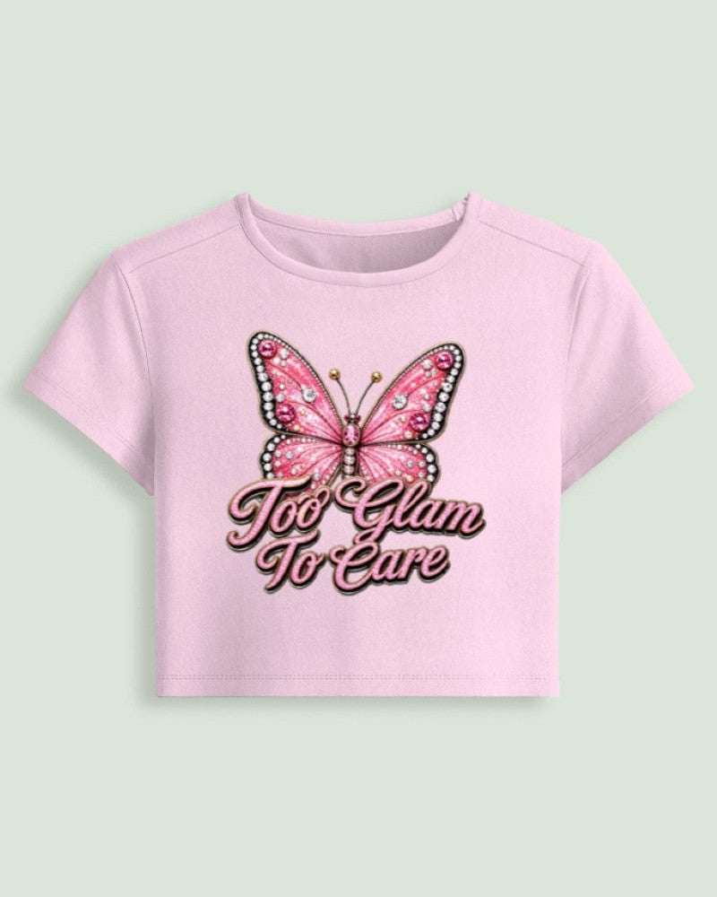 A light pink short-sleeve crop top featuring a graphic of a pink butterfly with rhinestone details and the text 'Too Glam To Care' in pink cursive, displayed on a solid pale green background.