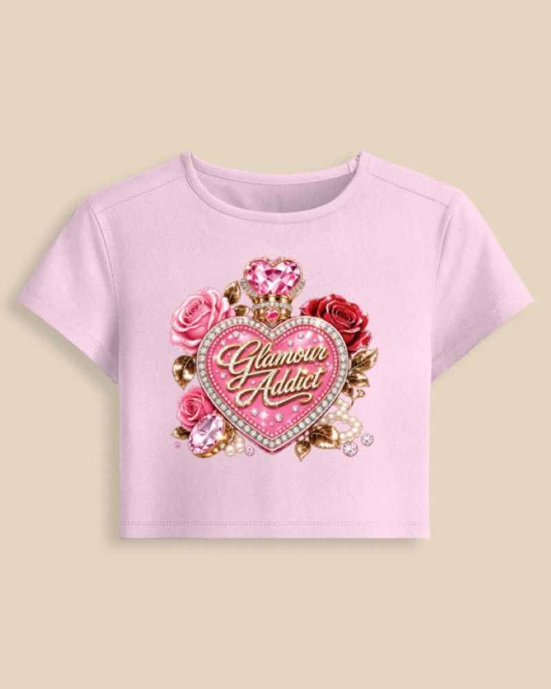 A light pink short-sleeve crop top featuring a large graphic on the front with a pink heart, roses, gemstones, and the words 'Glamour Addict' in gold script, shown against a solid beige background.