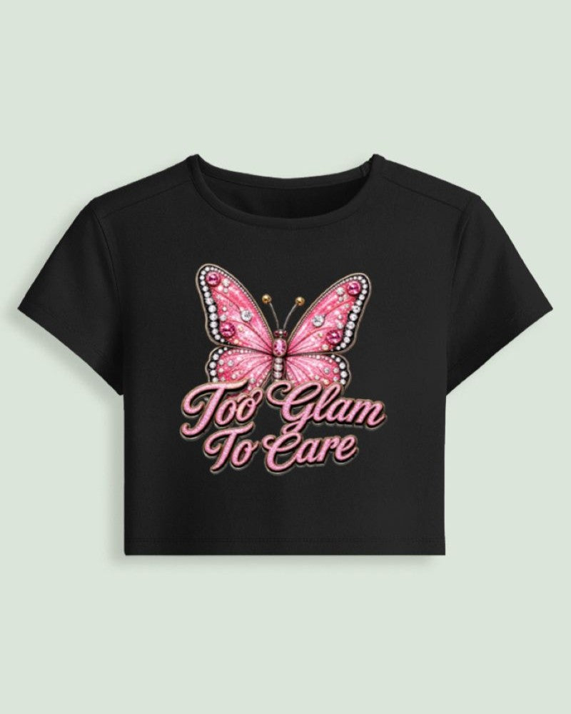 A black short-sleeve crop top t-shirt featuring a pink rhinestone-embellished butterfly graphic and the text 'Too Glam To Care' in pink script, displayed against a light sage green background.