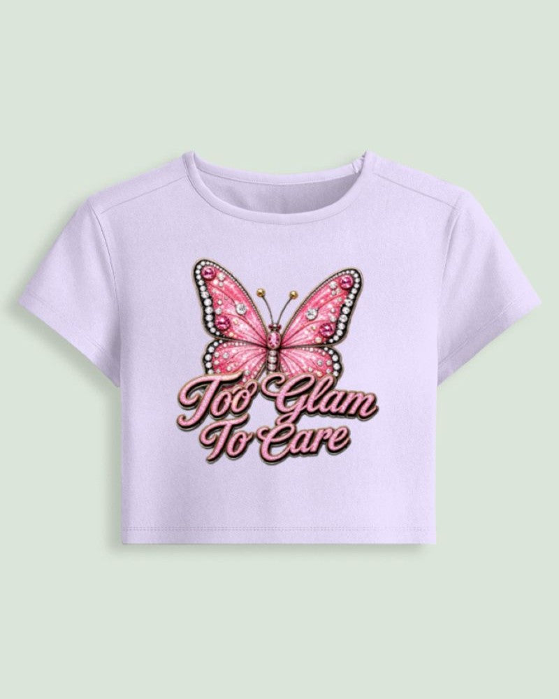 A light lavender short-sleeve crop top featuring a pink butterfly graphic and the text 'Too Glam To Care' in pink script on the front, displayed against a solid pale green background.