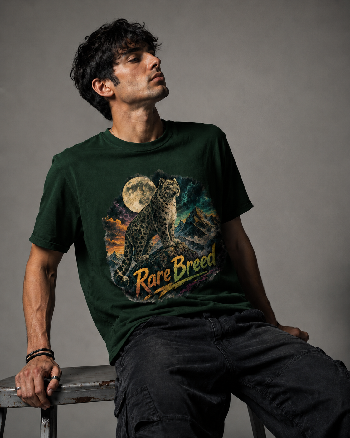 A man wearing a dark green graphic t-shirt with a rare breed cat print and the text 'Rare Breed' on the front, sitting on a stool against a gray studio background.
