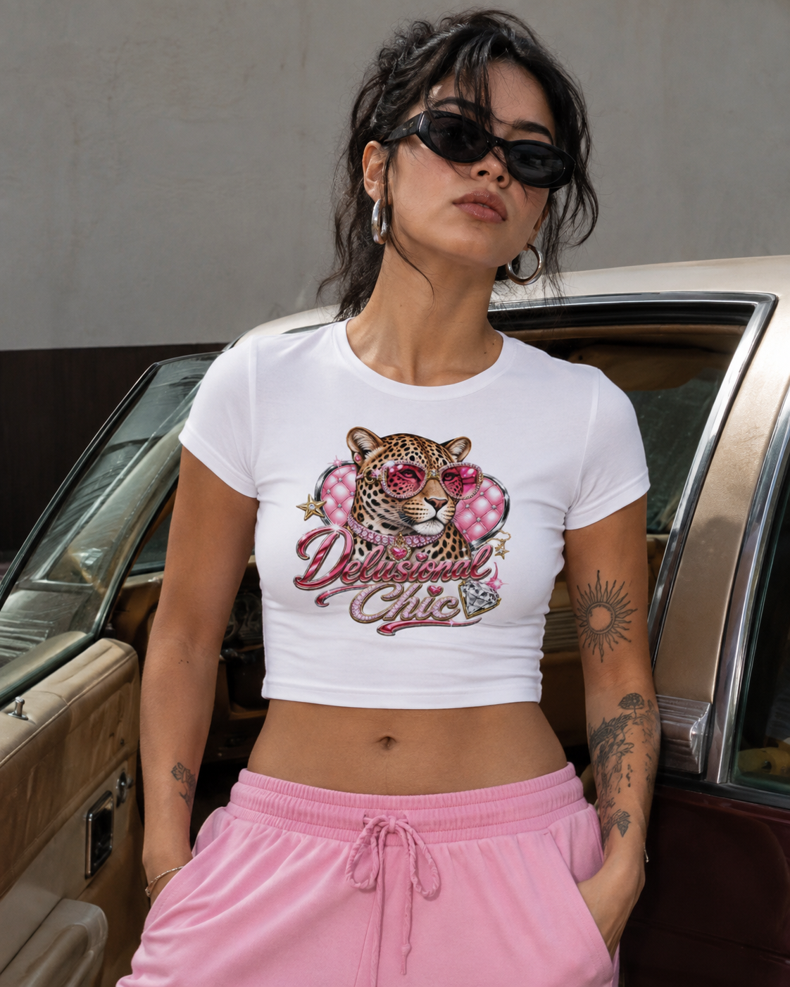 A woman wearing a white cropped t-shirt with a pink leopard graphic and the words 'Delicious Girls' in pink script, paired with pink sweatpants, standing next to a vintage car.