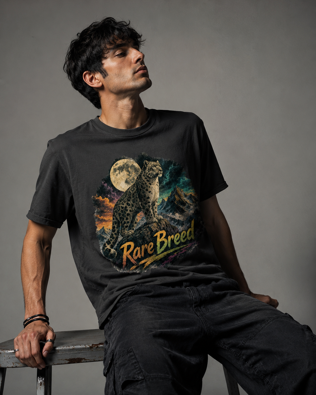 A man wearing a black graphic t-shirt with a detailed print of a leopard under a full moon and the text 'Rare Breed', sitting on a stool against a gray studio background.