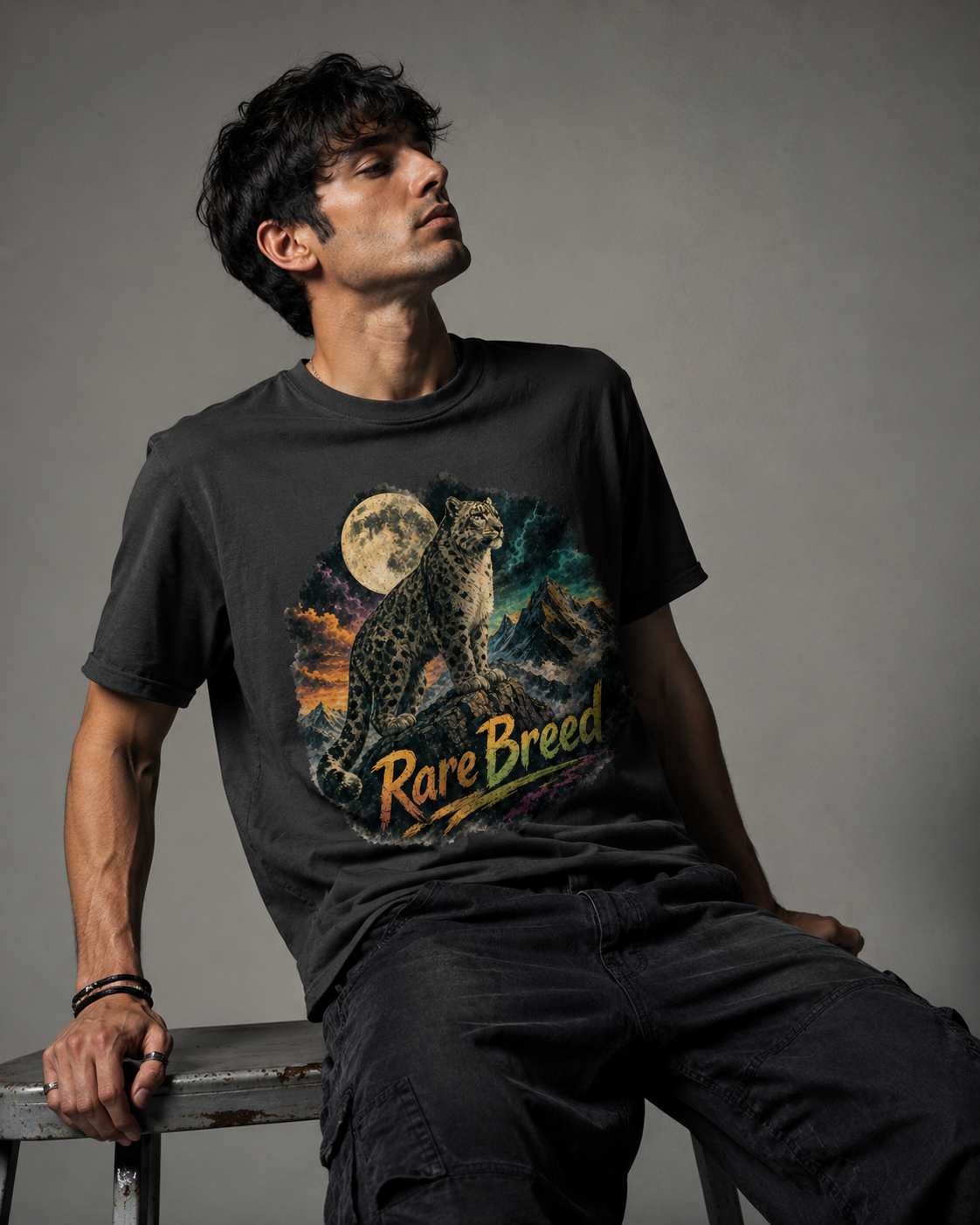 A man wearing a black graphic t-shirt with a detailed print of a leopard under a full moon and the text 'Rare Breed', sitting on a stool against a gray studio background.
