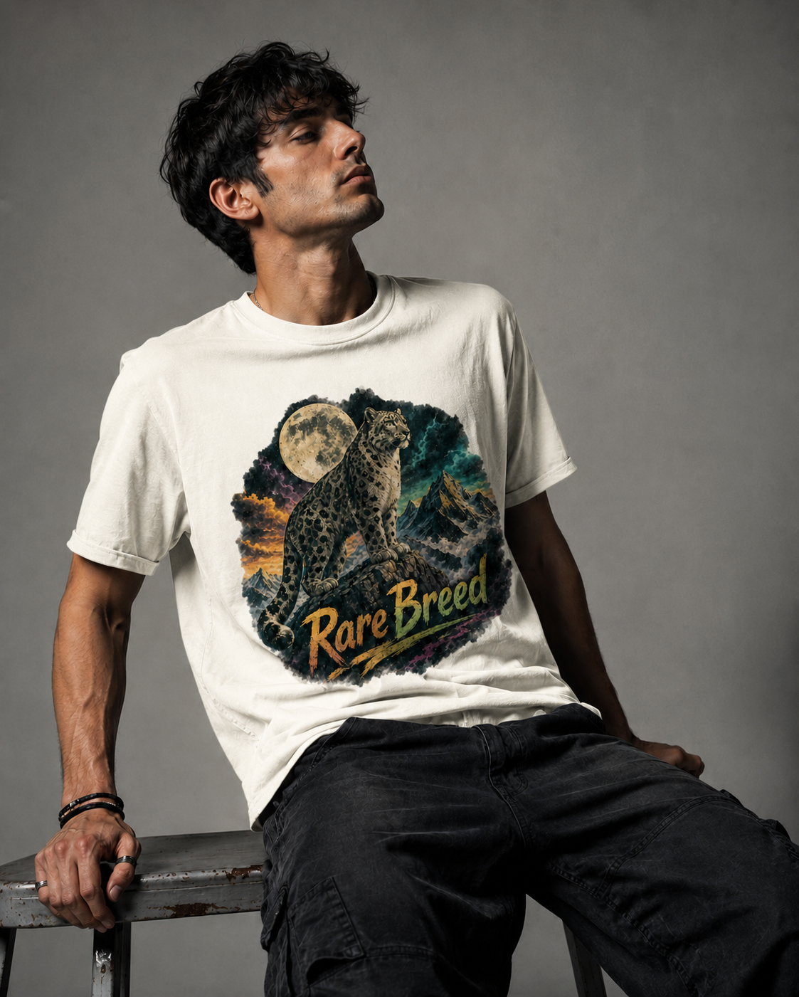 A man wearing a cream-colored graphic t-shirt with a detailed print of a cat and the words 'Rare Breed' on the front, paired with dark cargo pants, sitting on a stool against a gray studio background.