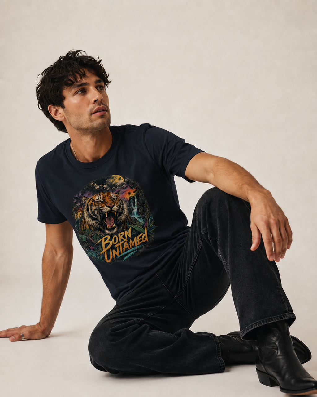 A man wearing a navy blue graphic t-shirt with a colorful tiger print and the text 'Born Untamed', paired with dark jeans and black boots, sitting against a light beige studio background.