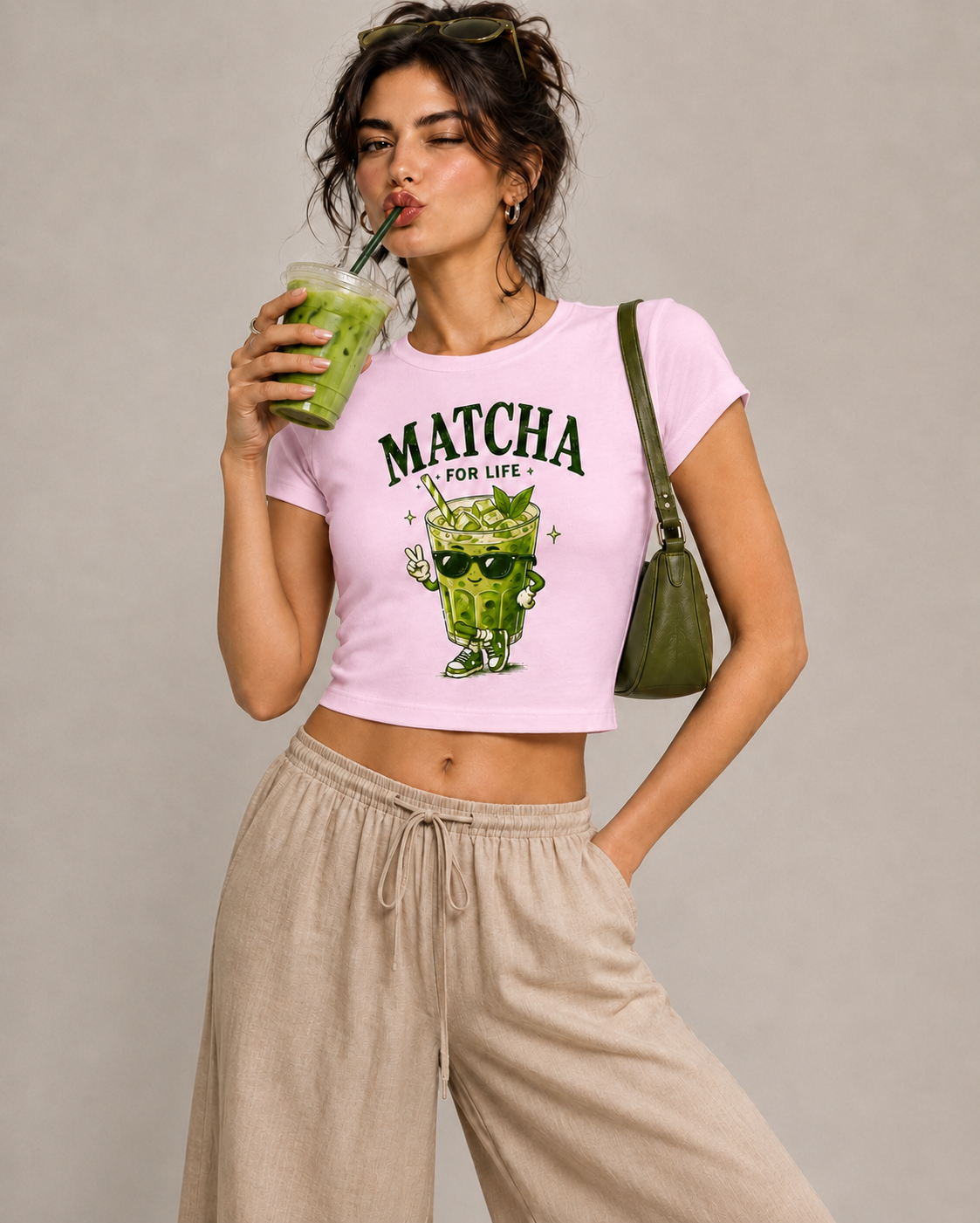 A woman modeling a light pink cropped t-shirt with a green matcha cartoon graphic and the text 'MATCHA FOR LIFE'. She is wearing beige drawstring pants, holding a green drink, and carrying an olive green shoulder bag against a neutral gray background.