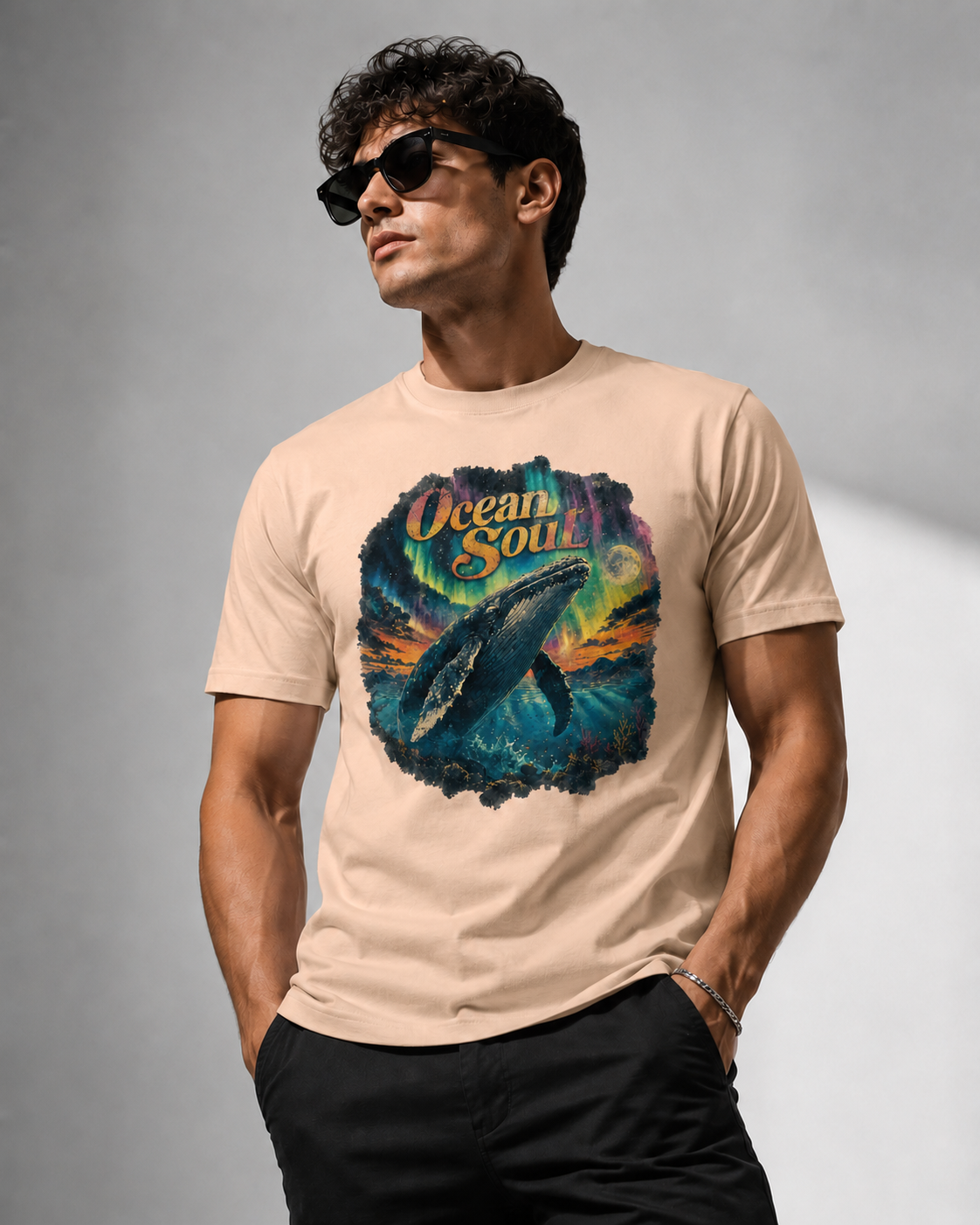 A man wearing a beige graphic t-shirt with a colorful ocean design and the words 'Ocean Soul', paired with black pants and sunglasses, set against a gray studio background.