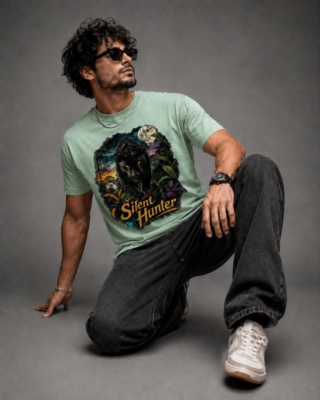 A man wearing a light green graphic t-shirt with a wolf design and the text 'Silent Hunter', paired with dark jeans and white sneakers, posing on a gray studio background.
