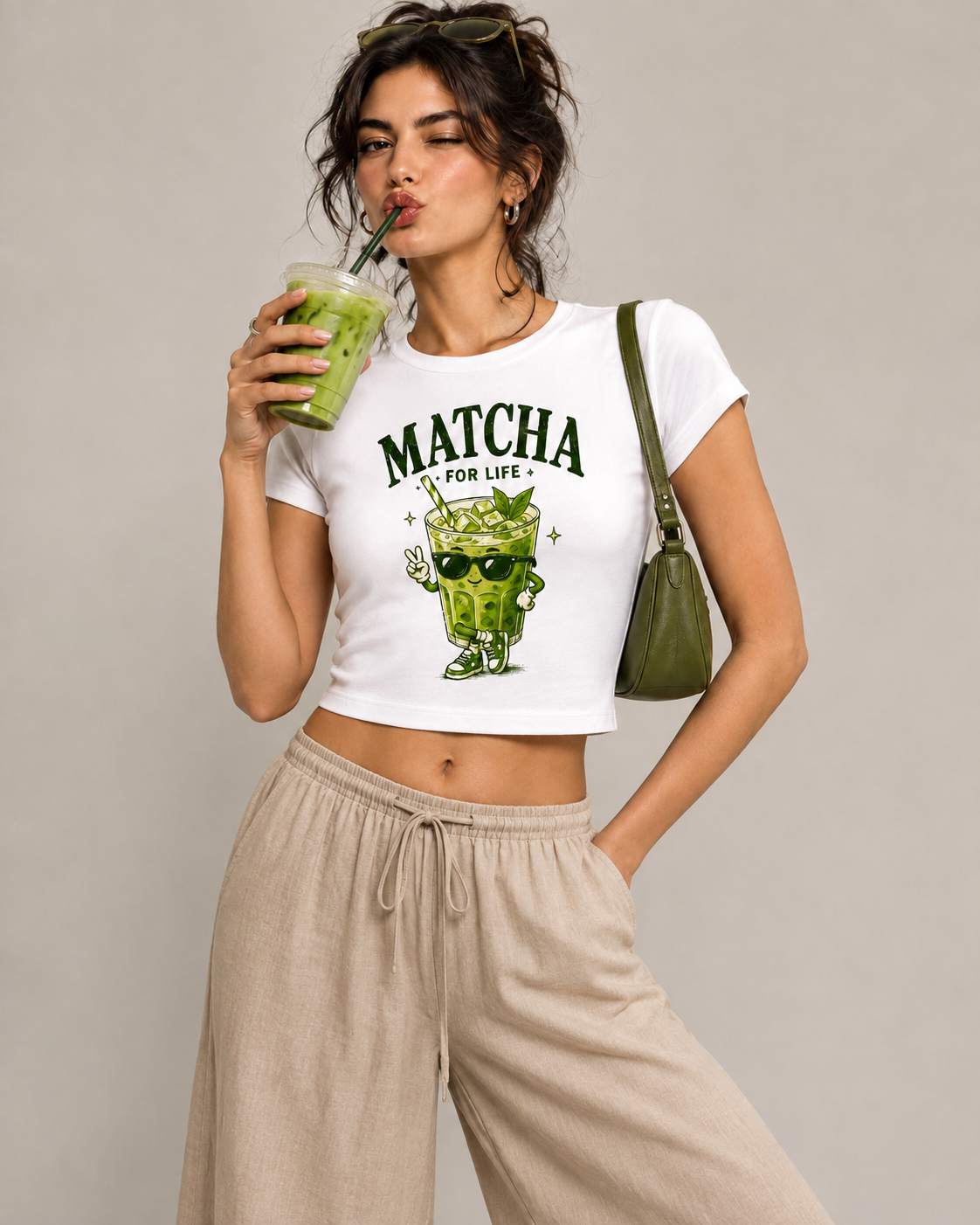 A woman modeling a white crop top t-shirt with a green matcha cartoon graphic and the text 'MATCHA FOR LIFE'. She is wearing beige drawstring pants, holding a green drink, and carrying an olive green shoulder bag against a solid light gray background.