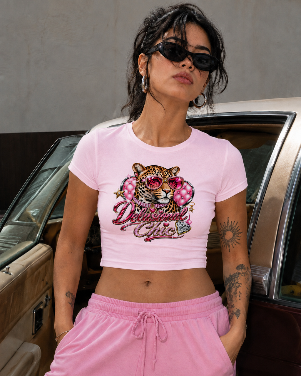 A woman wearing a light pink cropped t-shirt with a graphic of a leopard wearing pink sunglasses and the text 'Delilah Girls' in pink script, paired with pink sweatpants, posing in front of a vintage car.
