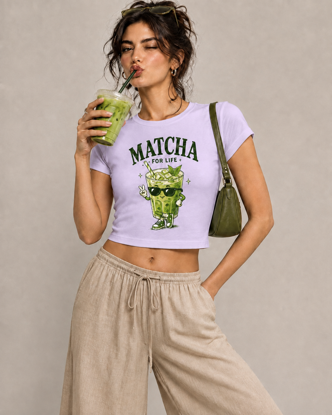 A woman modeling a light purple cropped t-shirt with a green matcha cartoon graphic and the text 'MATCHA FOR LIFE'. She is wearing beige drawstring sweatpants, holding a green drink, and carrying an olive green shoulder bag against a neutral beige background.