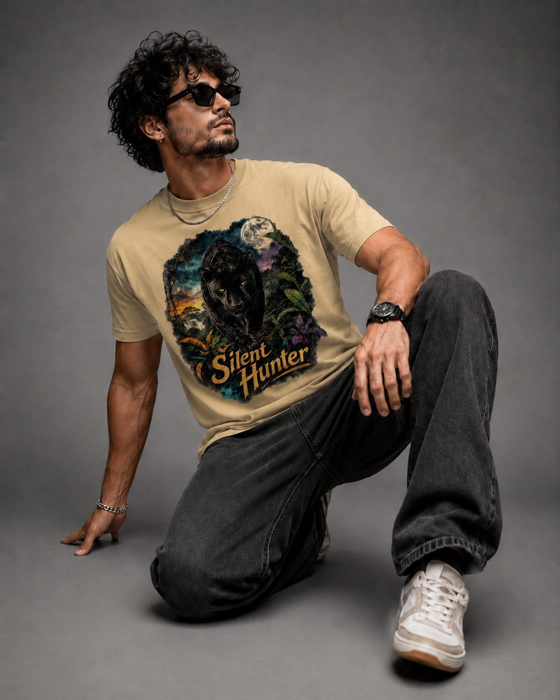 A man wearing a beige graphic t-shirt with a wolf design and the text 'Silent Hunter', paired with dark gray jeans and white sneakers, posing on a gray studio background.