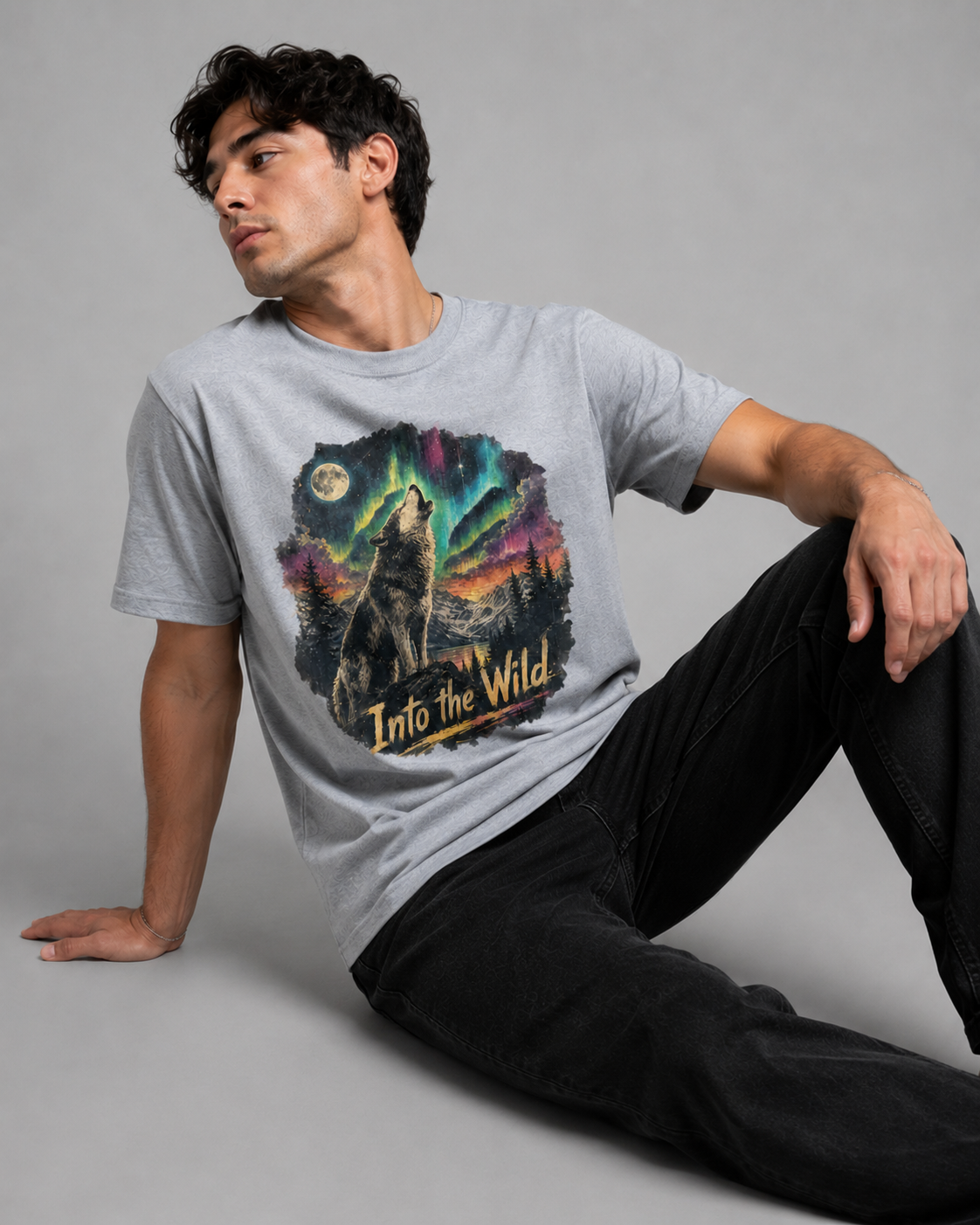 A man wearing a light gray heather graphic t-shirt with a wolf howling under a colorful aurora borealis night sky, paired with black jeans, posed against a solid gray studio background.