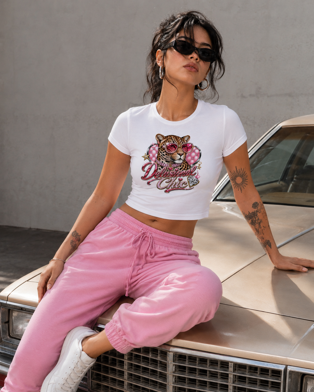 A woman wearing a white graphic crop top with a pink and gold cheetah design and the words 'Delilah Killer,' paired with pink sweatpants and white sneakers, sitting on the hood of a beige car against a gray wall.