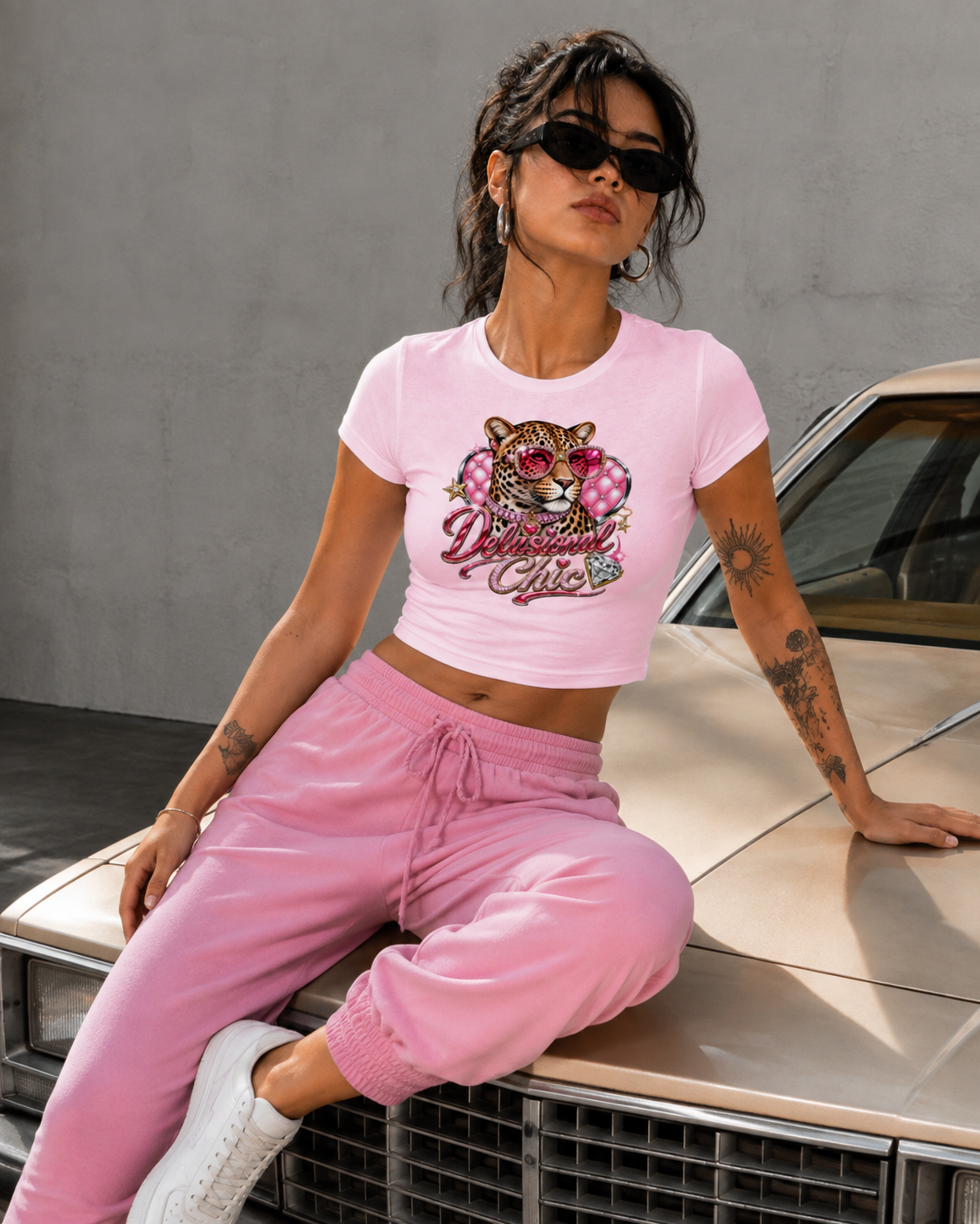 A woman wearing a light pink crop top with a 'Delicious Girl' tiger graphic and matching pink sweatpants, sitting on the hood of a beige car against a gray wall.