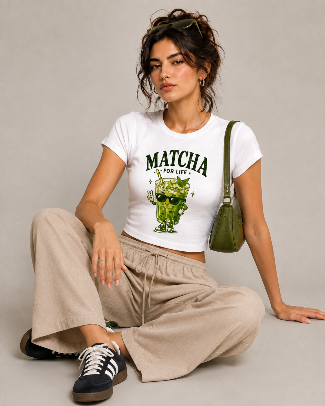 A woman sits cross-legged on a light gray studio floor wearing a white cropped T-shirt with a green 'MATCHA IS FOR LIFE' cartoon matcha graphic, beige wide-leg pants, black-and-white sneakers, and a green shoulder bag.