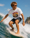 A man surfing on a wave wearing a white graphic t-shirt that reads 'Ride The Waves' in red and yellow, paired with dark shorts and sunglasses.