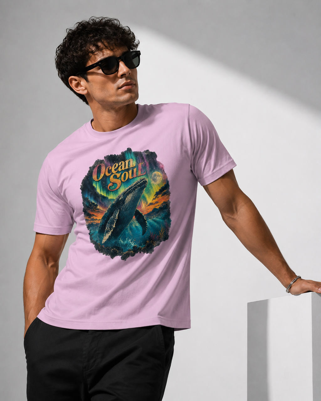 A man wearing a light pink t-shirt with a colorful 'Ocean Soul' whale graphic, styled with black sunglasses and black pants against a gray studio background.