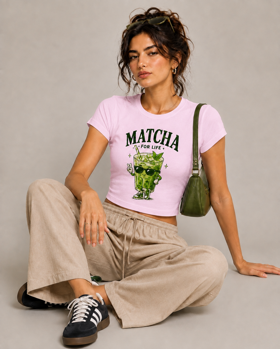 A woman sits on a studio floor wearing a light pink cropped T-shirt with a green matcha cartoon graphic and the text 'MATCHA FOR LIFE'. She is also wearing beige wide-leg pants, black and white sneakers, and carrying an olive green shoulder bag against a neutral gray background.