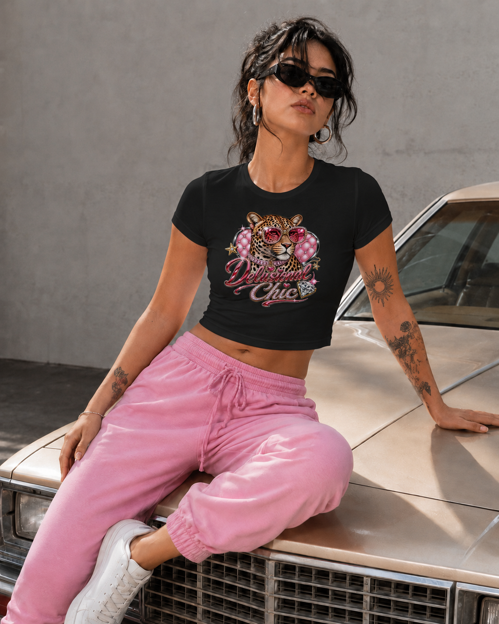 A woman wearing a black graphic crop top with a tiger and floral design, pink sweatpants, and white sneakers, sitting on the hood of a vintage car against a gray wall.