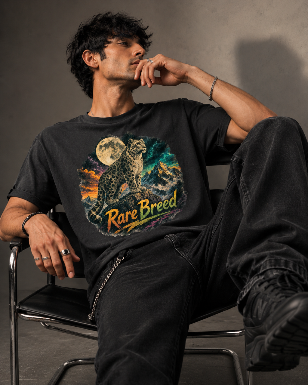A man wearing a black graphic t-shirt with a colorful print of a rare breed leopard under a full moon and the text 'Rare Breed', paired with black jeans, sitting on a chair in a studio setting.