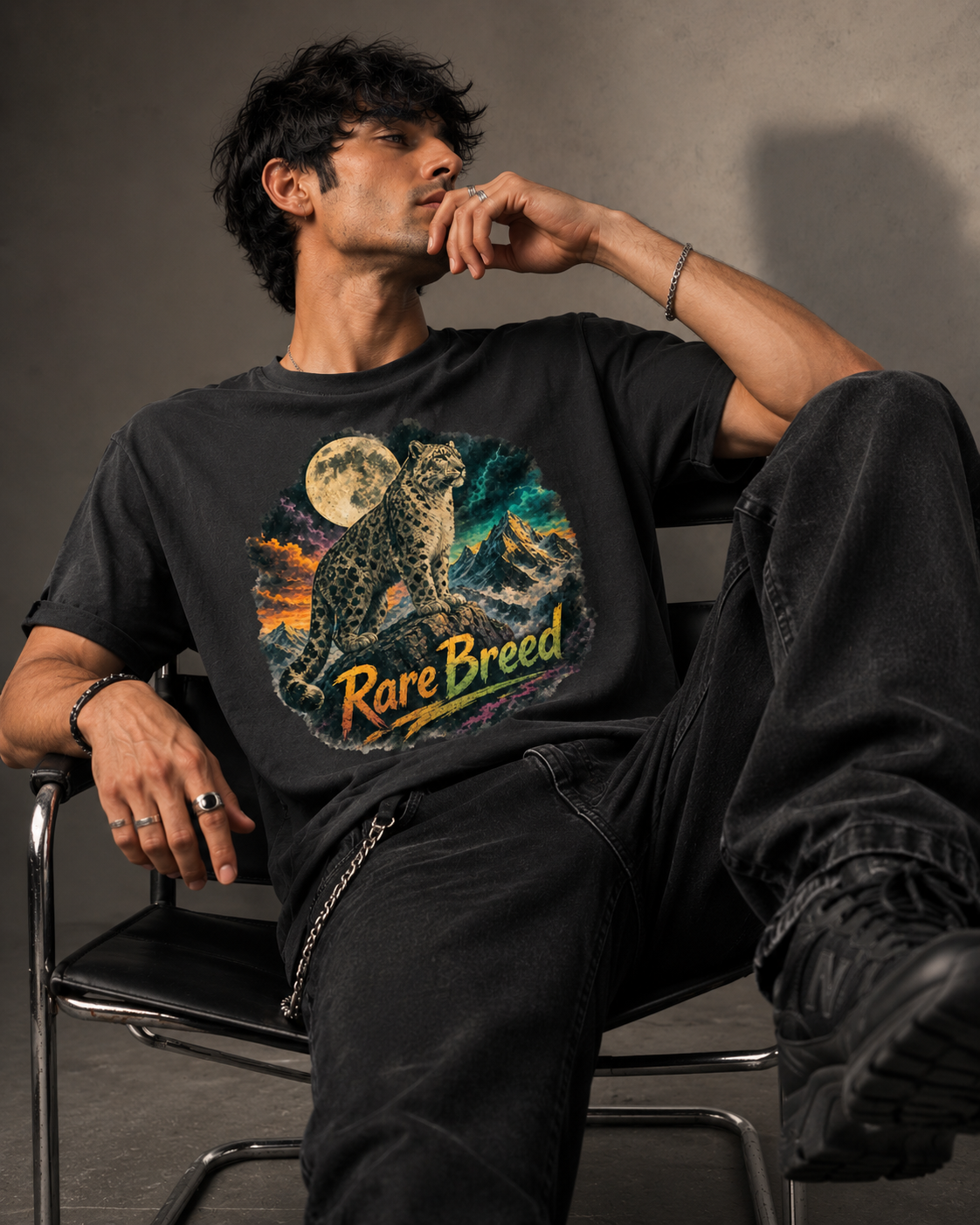 A man wearing a black graphic t-shirt with a colorful print of a rare breed leopard under a full moon and the text 'Rare Breed', paired with black jeans, sitting on a chair in a studio setting.