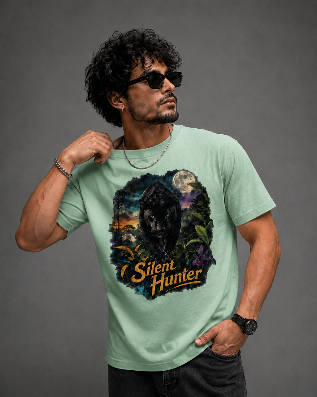 A man wearing a mint green graphic t-shirt with a 'Silent Hunter' black panther design, styled with black sunglasses, a silver chain necklace, and a black watch, set against a dark gray studio background.