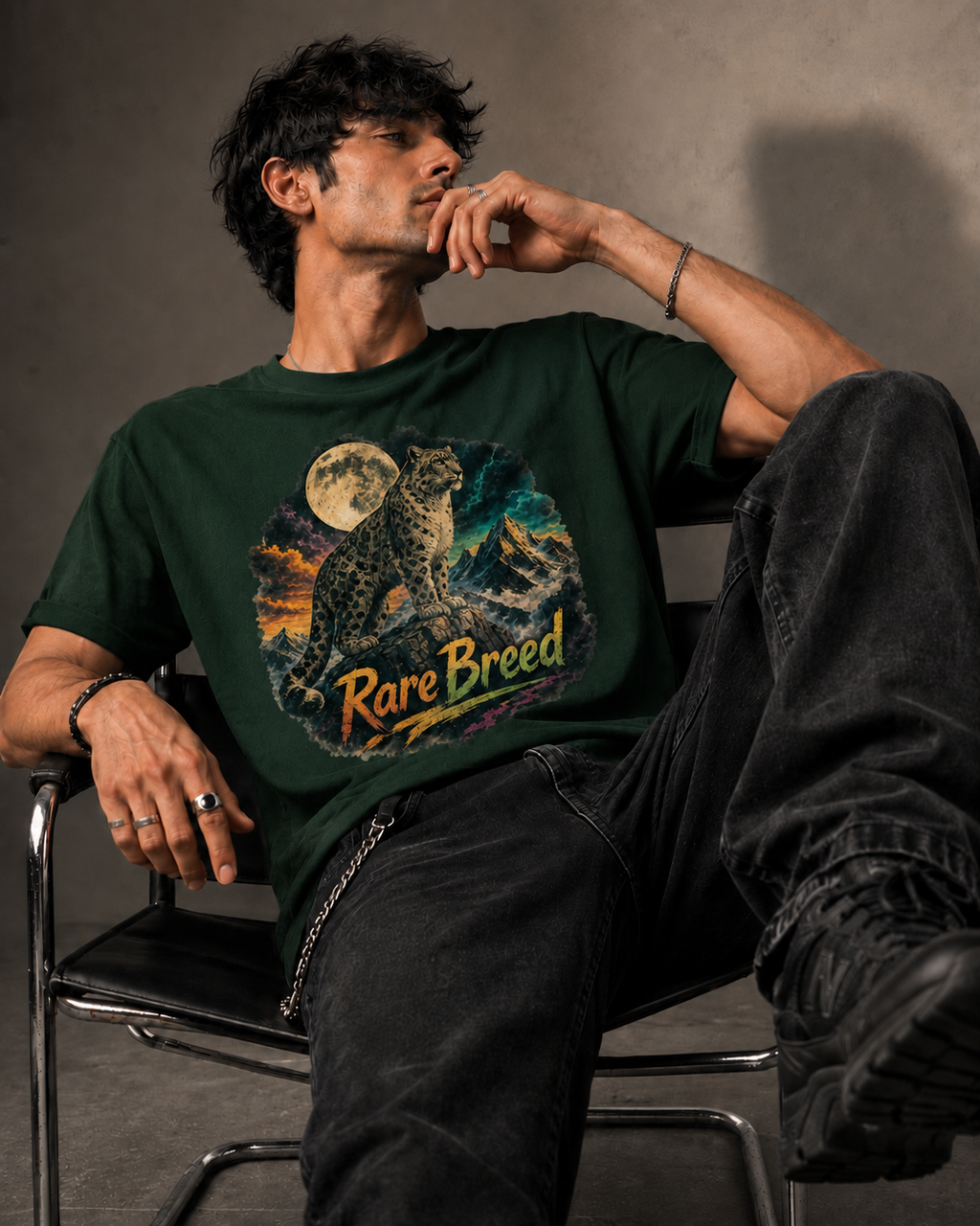 A man wearing a dark green graphic t-shirt with a detailed print of a leopard and the text 'Rare Breed', sitting on a black chair against a textured gray wall.