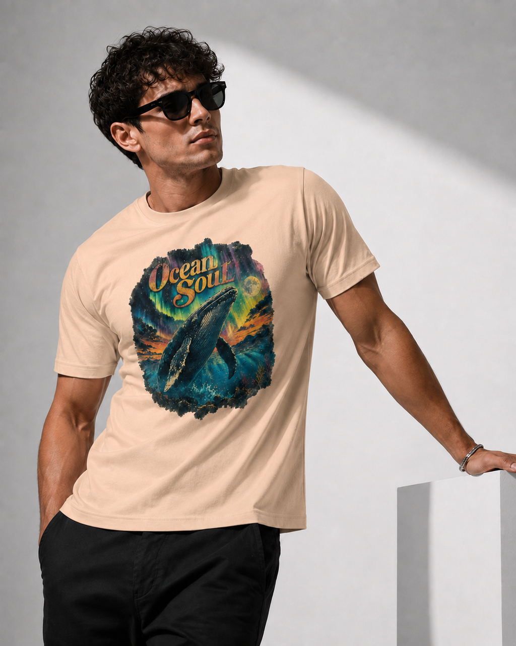 A man wearing a beige graphic t-shirt with a colorful ocean whale print and the words 'Ocean Soul', paired with black pants and sunglasses, standing against a gray wall.