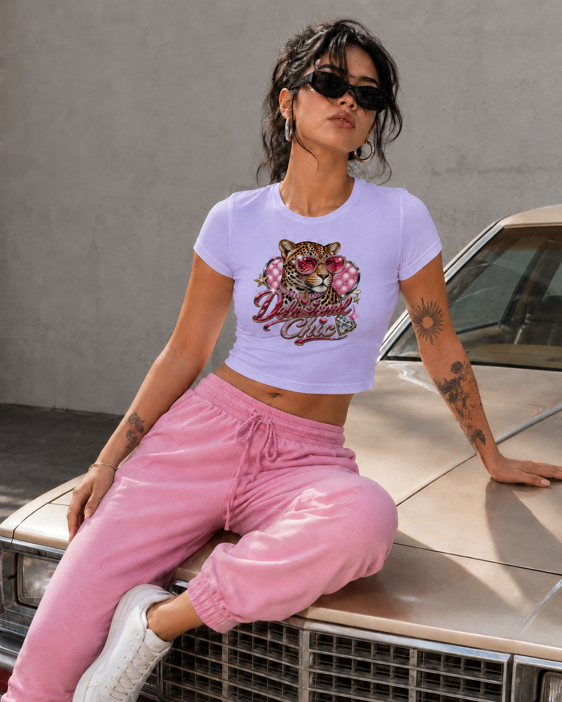 A woman wearing a light lavender crop top with a colorful tiger graphic and pink sweatpants, sitting on the hood of a vintage car against a gray concrete wall.