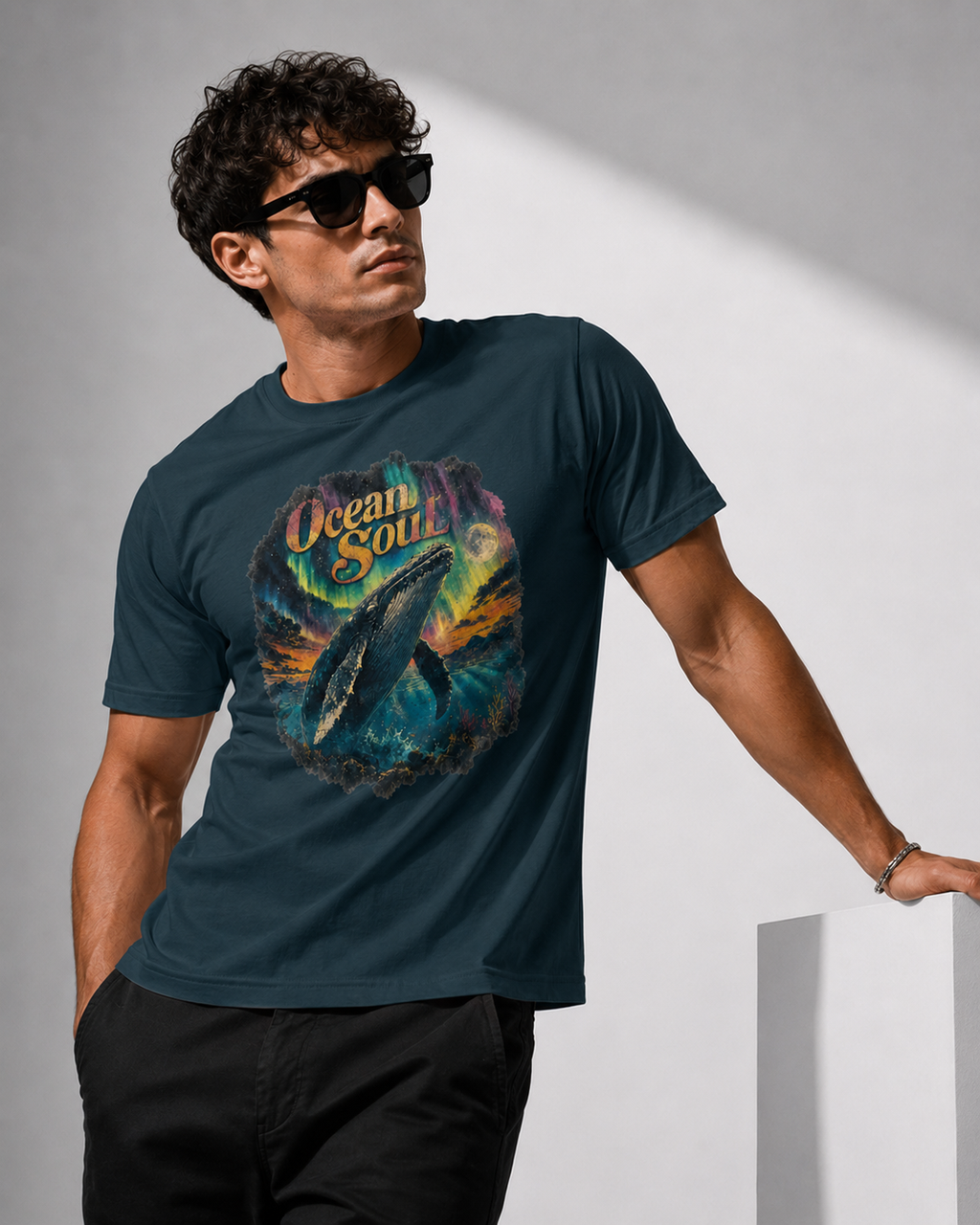 A man wearing a dark teal graphic t-shirt with a colorful ocean whale print and the words 'Ocean Soul', paired with black pants and sunglasses, standing against a minimalist gray background.