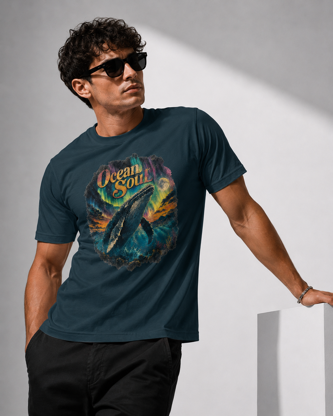 A man wearing a dark teal graphic t-shirt with a colorful ocean whale print and the words 'Ocean Soul', paired with black pants and sunglasses, standing against a minimalist gray background.