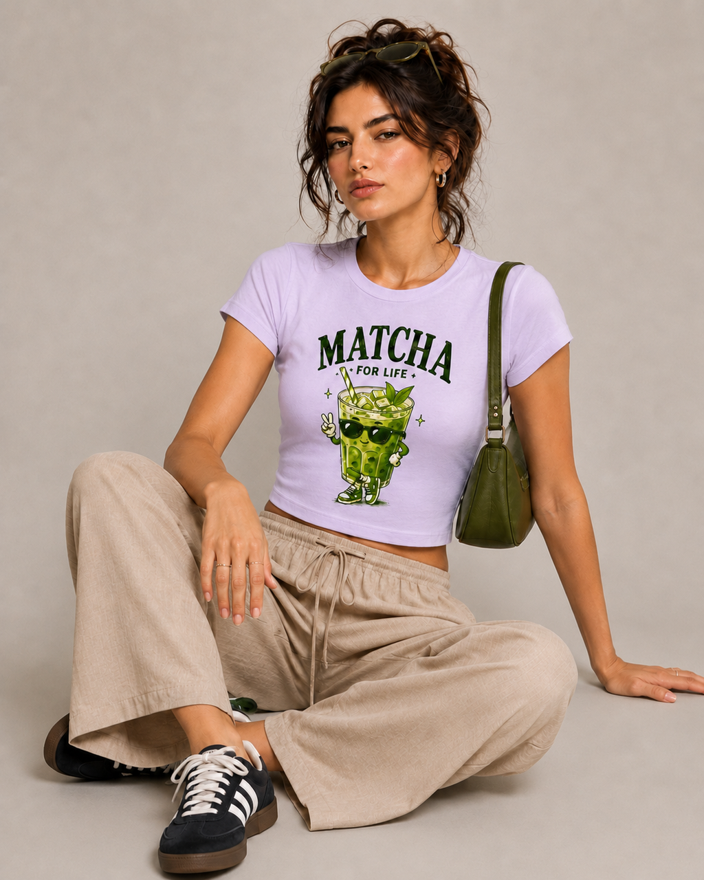 A woman sits cross-legged on a neutral studio floor wearing a light lavender cropped T-shirt with a green cartoon matcha graphic and the text 'MATCHA IS FOR LIFE'. She is styled with beige wide-leg sweatpants, black and white sneakers, and a green shoulder bag.