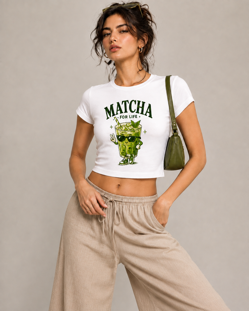 A woman modeling a white cropped t-shirt with a green 'MATCHA FOR LIFE' cartoon graphic, paired with beige wide-leg sweatpants and a green shoulder bag, against a neutral gray background.
