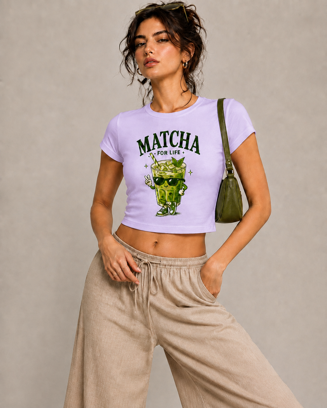 A woman modeling a light purple cropped t-shirt with a green and black 'MATCHA FOR LIFE' cartoon graphic, paired with beige wide-leg pants and a green shoulder bag, against a neutral gray background.