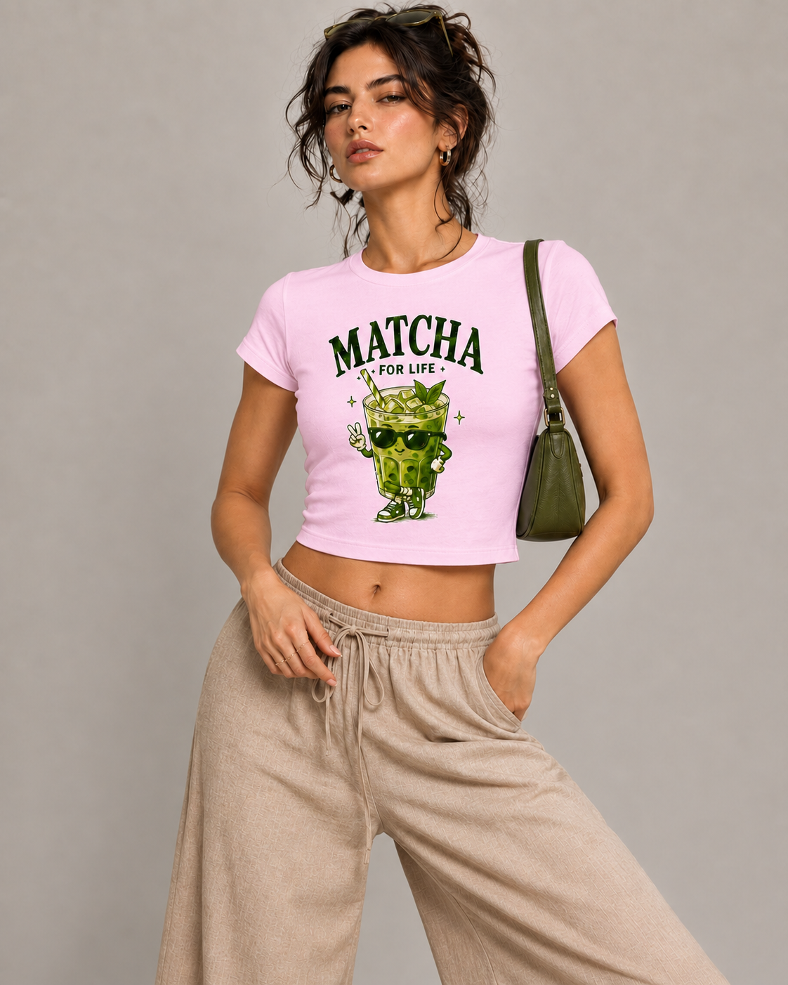 A woman modeling a light pink cropped t-shirt with a green matcha drink graphic and the text 'MATCHA FOR LIFE'. She is wearing beige wide-leg sweatpants and carrying an olive green shoulder bag against a solid gray studio background.